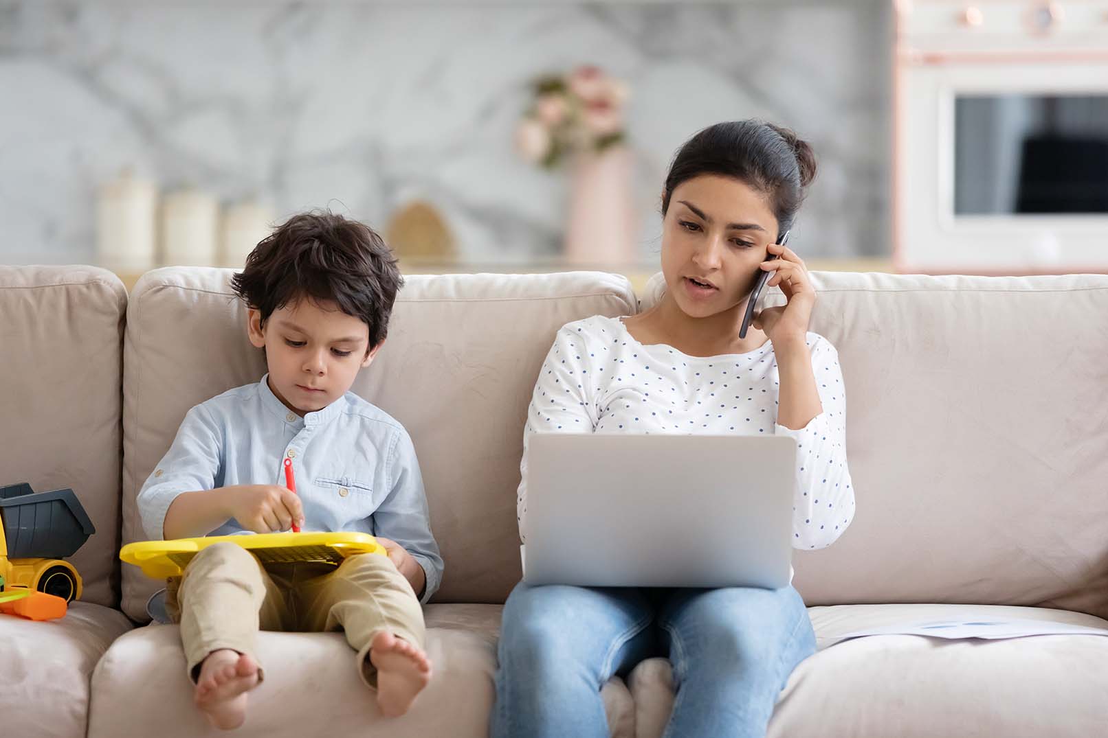 Mulher sentada no sofá de casa falando no telefone e usando o computador, com o filho desenhando ao lado.